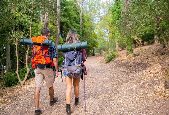 Back view of couple going along road in forest. Long-haired woman and man carrying backpacks and hiking on nature together. Green trees on background. Tourism, adventure and summer vacation concept